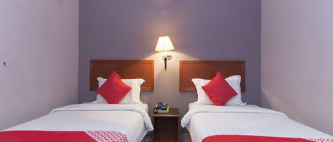 A hotel room with two single beds, each with a red and white bedspread, a wooden headboard, a bedside table with a lamp, and an air conditioning unit on the wall.