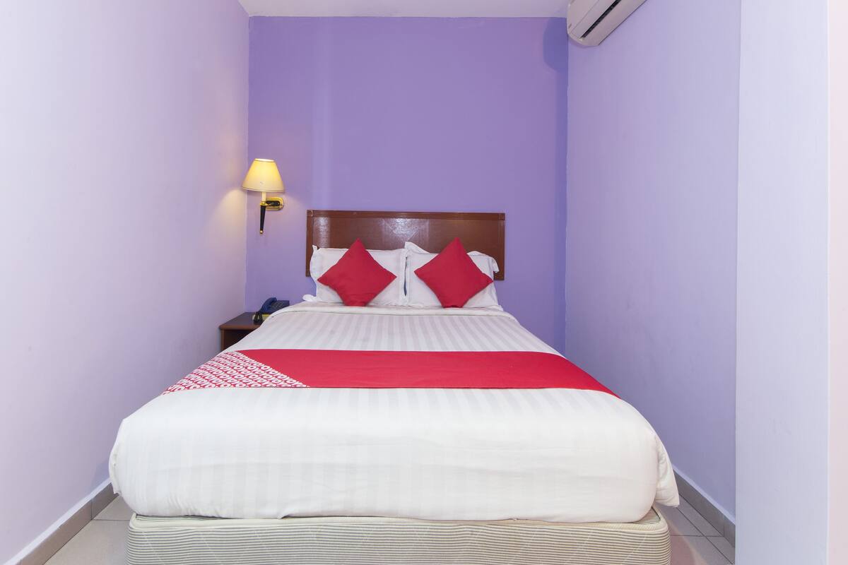 A hotel room with a bed, a nightstand, a lamp, and an air conditioner.