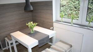 Una cocina moderna con encimera blanca, una mesita y un banco. Hay tres jarrones con plantas en el alféizar de la ventana.