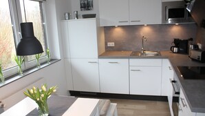 Una cocina moderna con armarios blancos, un lavamanos de acero inoxidable y un microondas. Hay una ventana con una lámpara colgante negra y un florero con flores en la encimera.