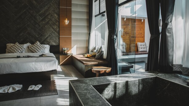 A modern hotel room with a large bed, a wooden bench, a small table, and a view of the outdoors through large windows. - Arun Dara Villa (Chiang Mai)