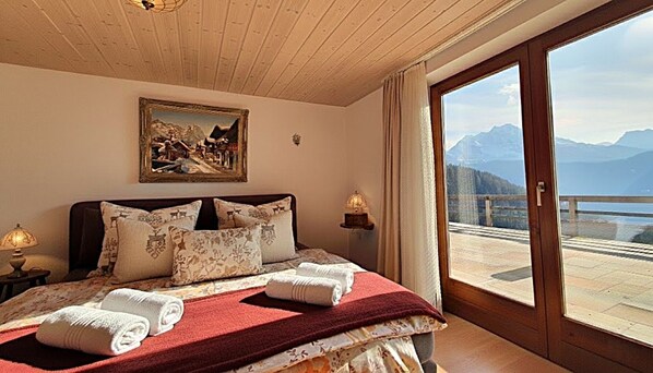 A bedroom with a large bed, a painting above the bed, and a view of mountains through a glass door.