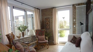 A cozy living room with a white sofa, wicker chairs, a small table with a lamp, and a view of the outdoors through large windows.