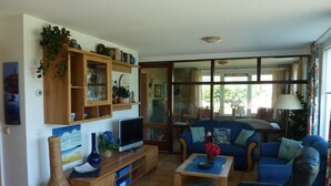 A living room with a television, sofa, and coffee table. There is a bookshelf with various items and a window with curtains.