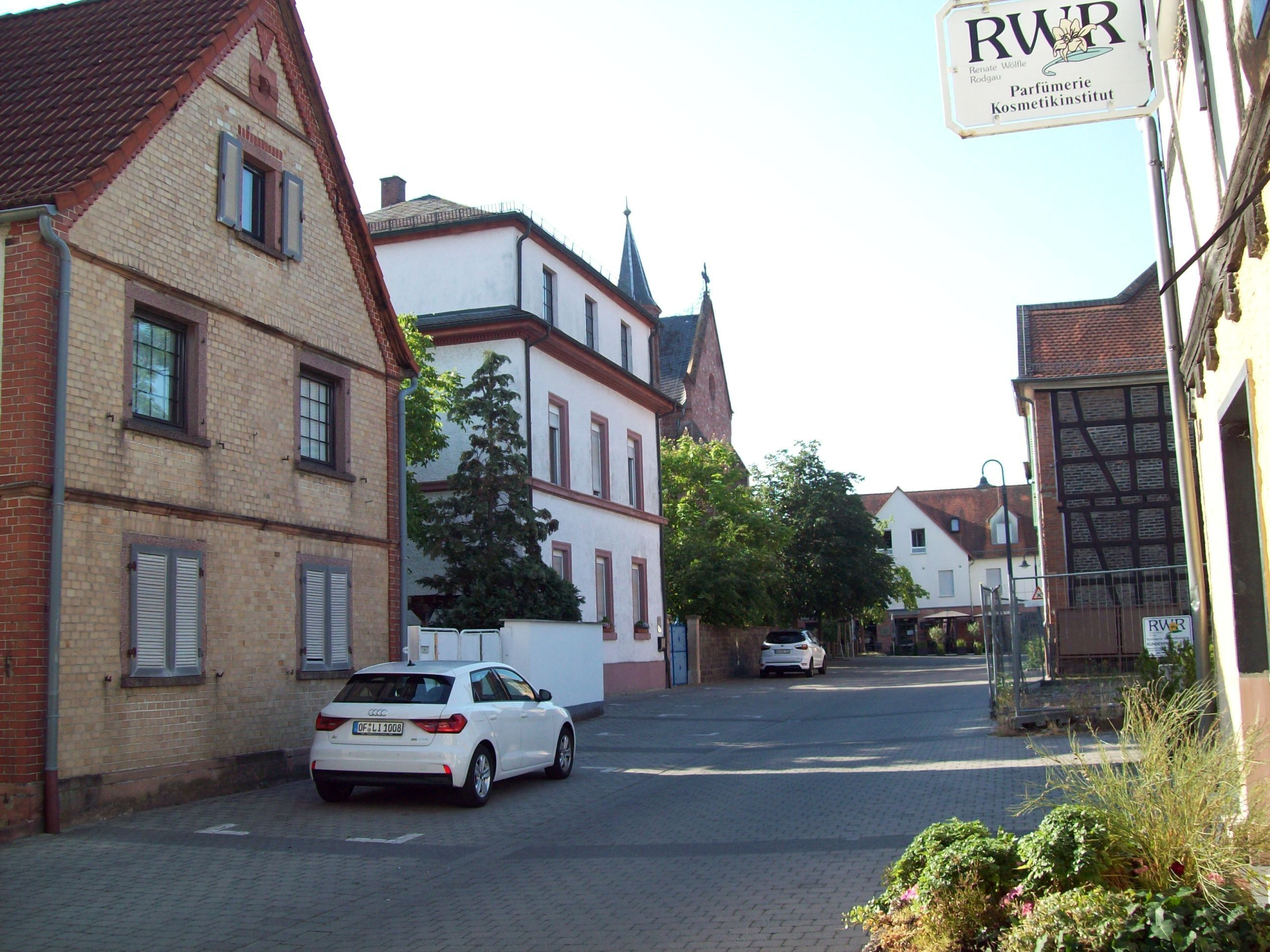 Una calle con arquitectura europea tradicional, un coche blanco estacionado al lado y un cartel para un instituto de perfumería.