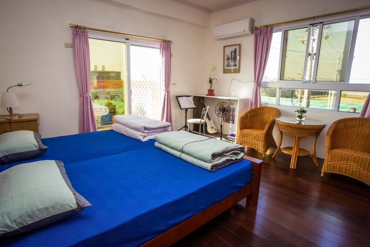 Une chambre avec un grand lit, deux chaises, un bureau avec un ventilateur et une fenêtre avec des rideaux.