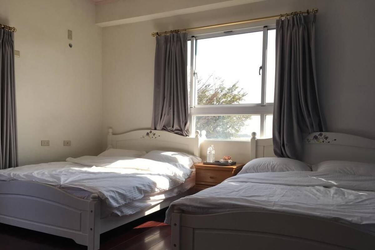 Une chambre avec deux lits simples, une fenêtre avec des rideaux, une table de chevet avec une bouteille d’eau et une petite commode.