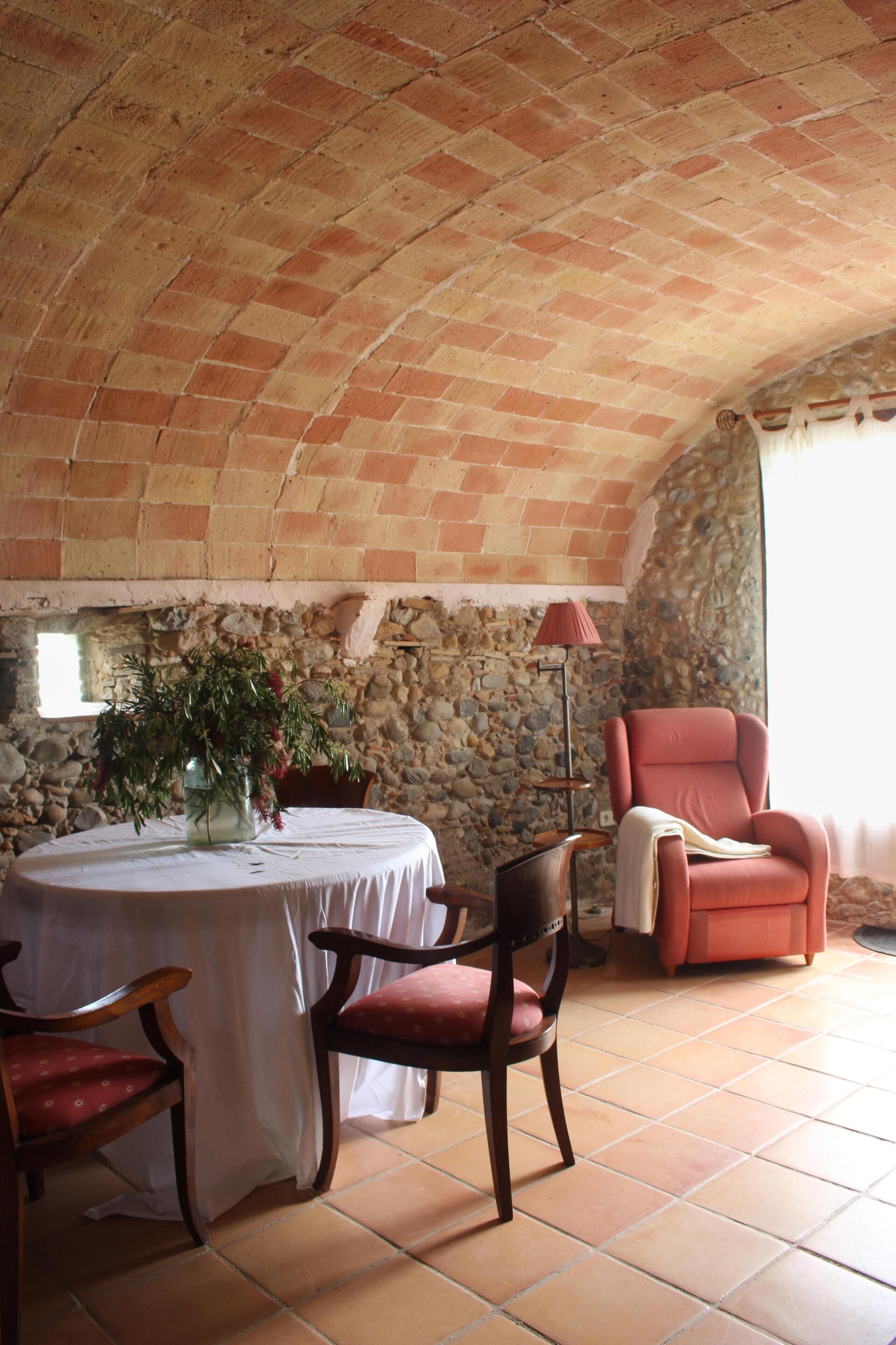 Une pièce avec une table ronde, deux chaises en bois, un fauteuil rouge, un vase avec des plantes vertes, une lampe et un mur en pierre.