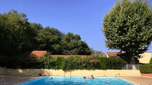Une piscine d’un bleu éclatant, entourée d’une terrasse dallée, où quelques personnes se baignent, et un arbre qui offre de l’ombre.