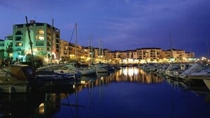 Un port de plaisance au crépuscule, avec des bateaux amarrés, des bâtiments dotés de balcons et une surface d’eau calme qui reflète les lumières.
