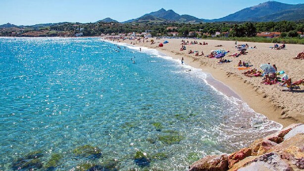 Une plage avec une eau d’un bleu limpide, un rivage sablonneux et des montagnes en arrière-plan.