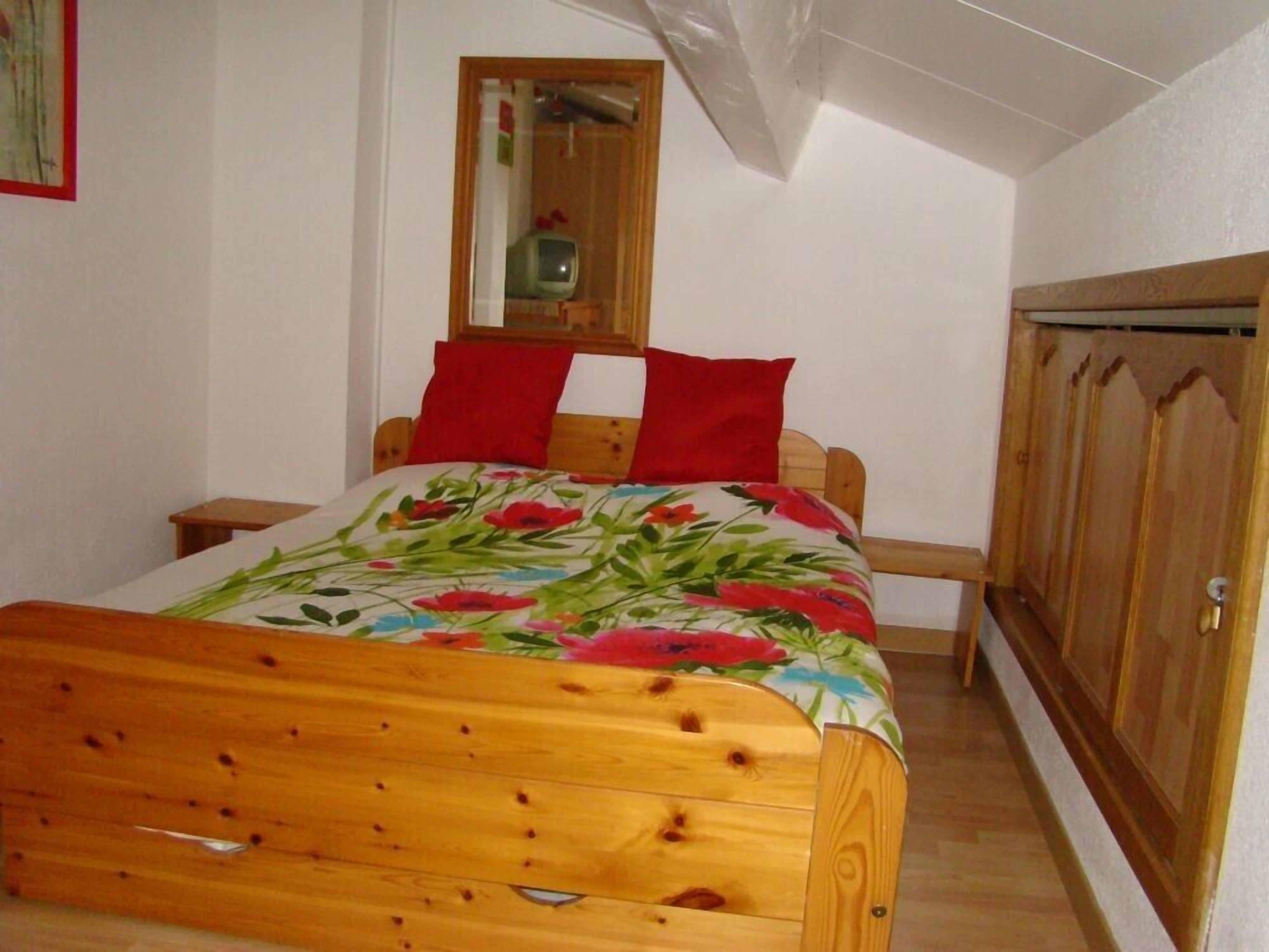 Une chambre avec un lit en bois, des oreillers rouges, une couvre-lit à motifs floraux, une armoire en bois et un miroir au mur.