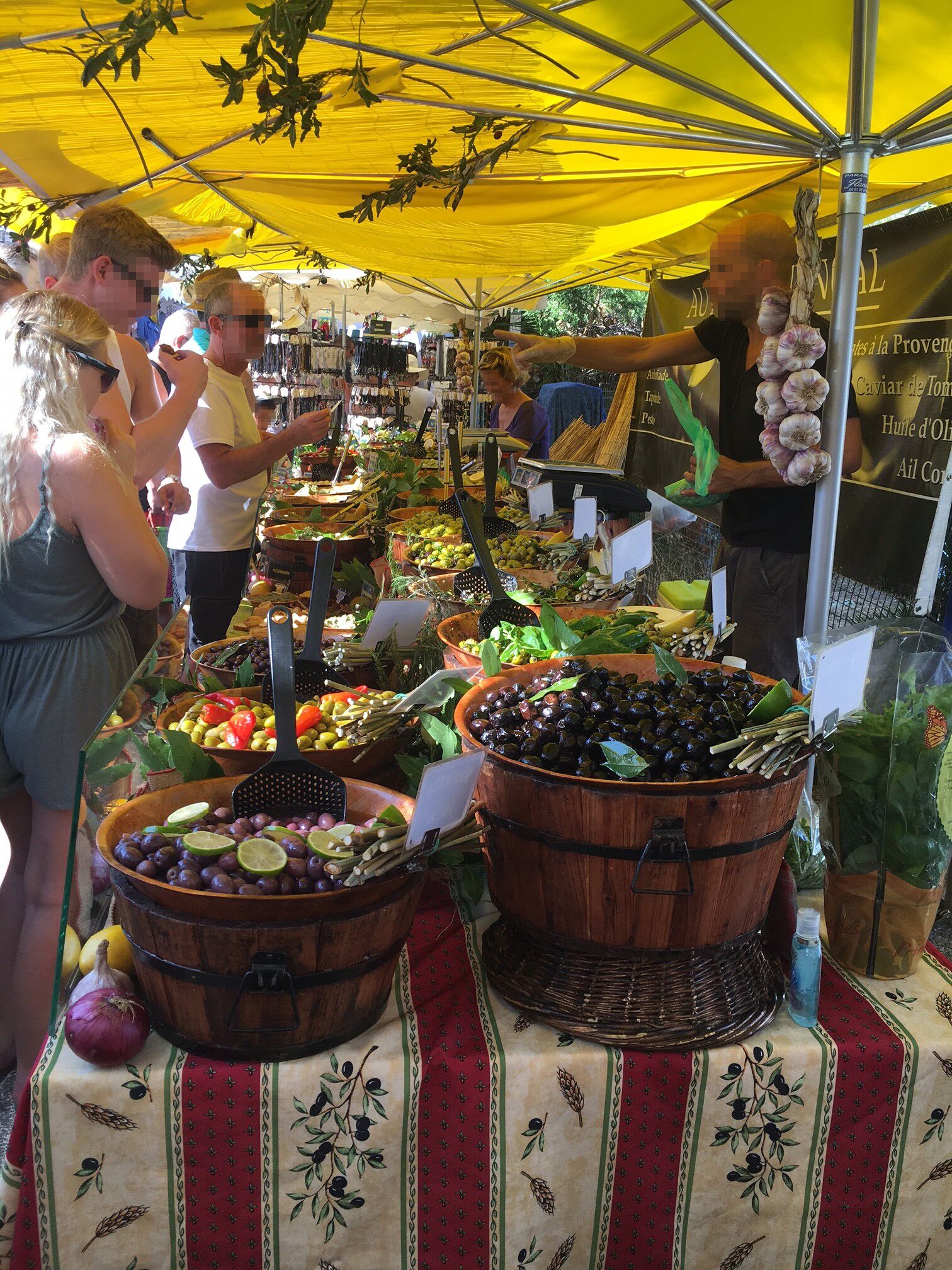 Un étal de marché proposant une variété de légumes et de fruits, notamment des olives et des oignons, sous un auvent jaune.