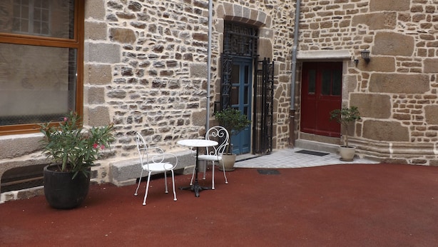 Un bâtiment en pierre avec une porte rouge, une porte bleue et une fenêtre. On y trouve deux plantes en pot et une table avec des chaises à l’extérieur.