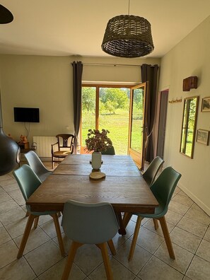 Un comedor con una mesa de madera, seis sillas, una lámpara colgante de fibras tejidas, un televisor en la pared y vistas a un jardín a través de un ventanal.