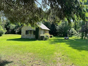 Un rustico cottage in pietra con una piccola finestra, immerso in un prato erboso e circondato da alberi.