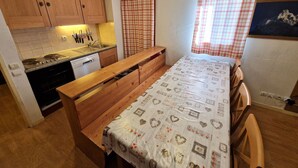 A compact kitchen with wooden cabinets, a stove, and a sink. There is a small dining area with a bench and a table covered with a patterned tablecloth. - 4-room apartment, 6-8 people, near ski slopes (Valfréjus)