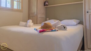 Ein ordentlich gemachtes Bett mit weißen Leinentüchern, einem zusammengerollten Handtuch und einem Strohhut auf dem Bett.