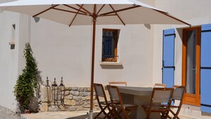 Um pátio externo com mesa e cadeiras, um guarda-sol branco e uma parede de pedra.