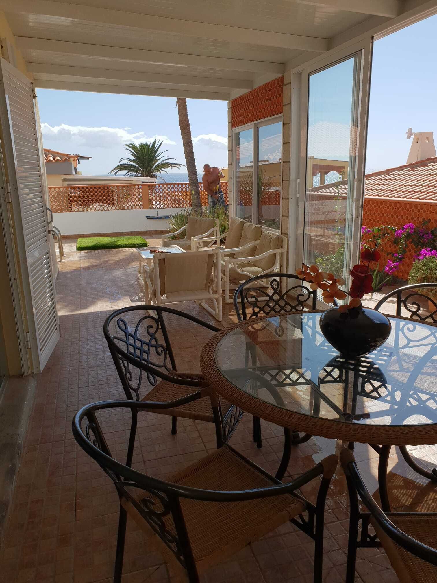 A covered patio with a glass table and chairs, a view of the sea, and a decorative vase.