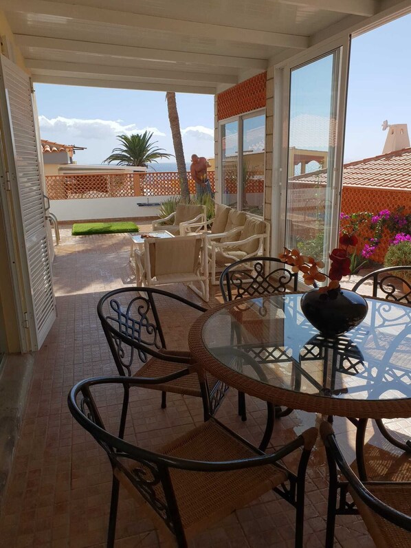 A covered patio with a glass table and chairs, a view of the sea, and a decorative vase.