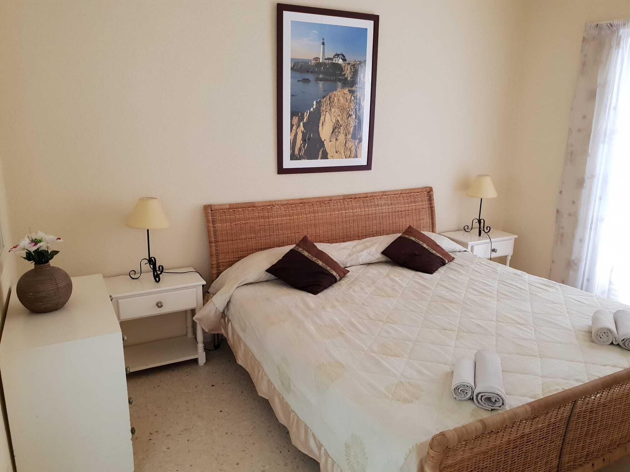 Ein ordentlich bezogenes Bett mit einer gesteppten weißen Decke, zwei braunen Kissen und auf einem weißen Nachttisch gerollten Handtüchern. An der Wand hängt ein gerahmtes Bild einer Küstenlandschaft.