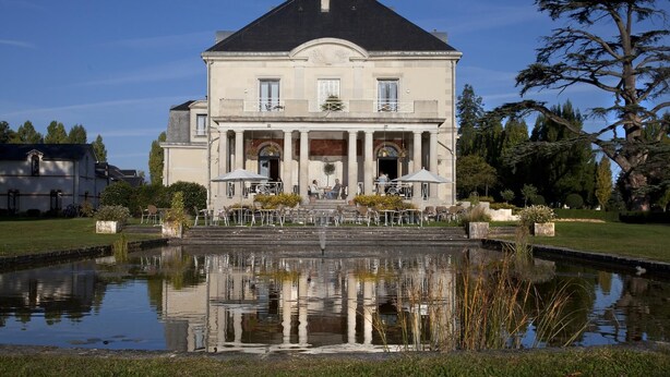 Um edifício grande e imponente com fachada simétrica, entrada central com colunas e um jardim ao redor com um lago refletor.