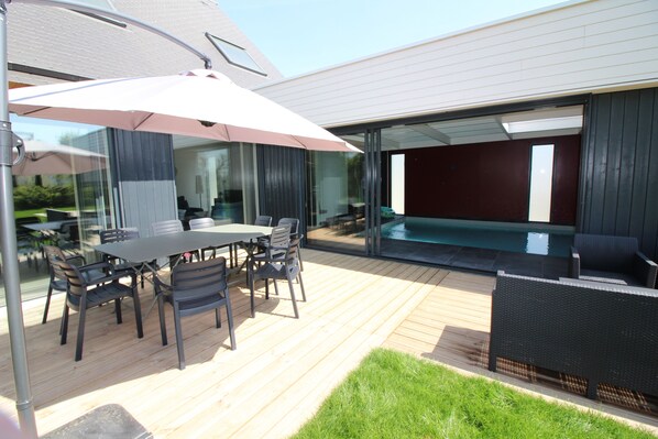 Un patio exterior moderno con una mesa de comedor y sillas, una sombrilla grande y una zona de piscina con paredes de vidrio.