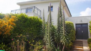Une maison moderne à deux étages, dotée d’un balcon, d’une porte d’entrée et d’un jardin bien entretenu où poussent différentes espèces de plantes.