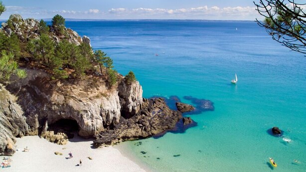 Une plage côtière aux eaux turquoise et limpides, une falaise rocheuse avec une entrée de grotte, et une plage de sable où l’on peut voir des gens et un voilier.