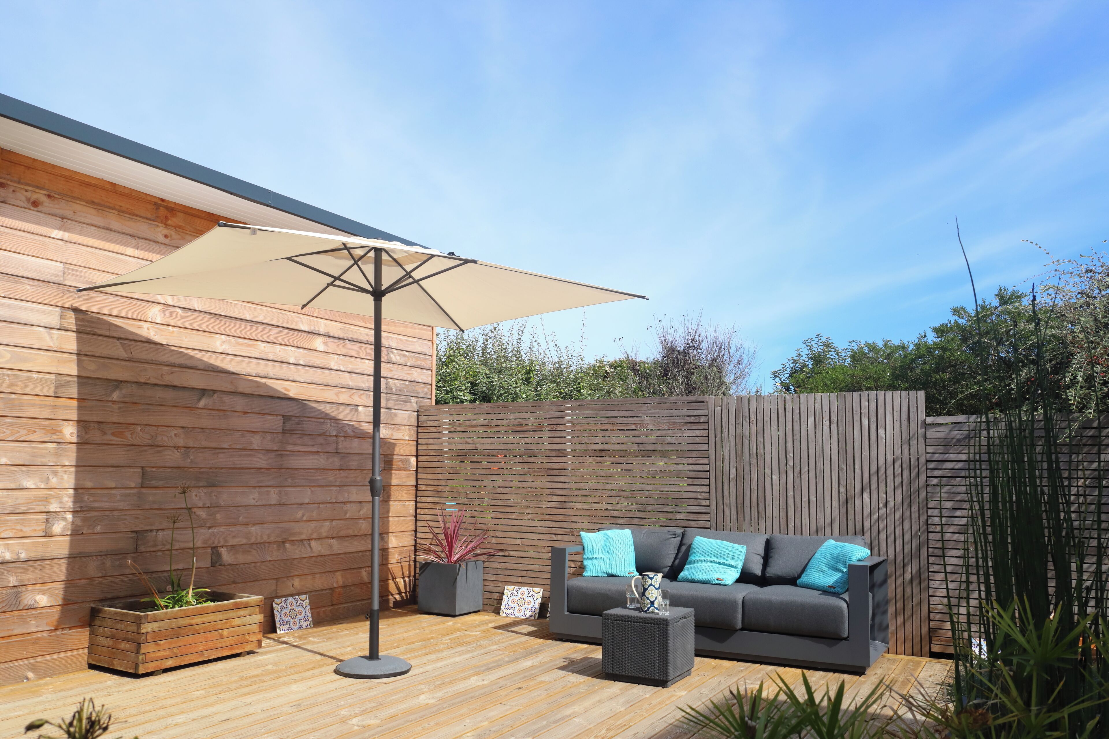 A modern outdoor seating area with a wooden deck, a sofa with blue cushions, and a large umbrella.