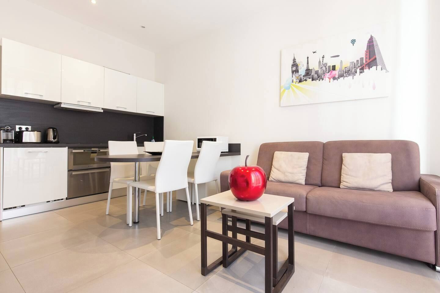 A modern kitchen with white cabinets, a dining area with white chairs, a brown sofa, and a red apple on a table.