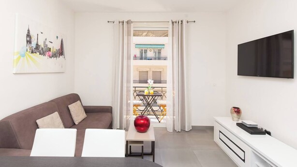 Ein modernes Wohnzimmer mit Sofa, Tisch mit einem roten Apfel, Fernseher und Blick auf einen Balkon.