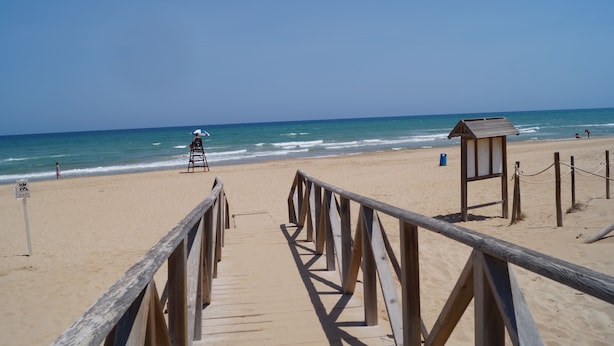Ein hölzerner Steg führt zu einem Strand mit einem Rettungsturm und einer kleinen Holzkonstruktion.