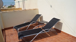 Eine Dachterrasse mit drei schwarzen Liegestühlen und einem kleinen Tisch, umgeben von einer weißen Mauer und einem gefliesten Boden.