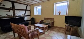 Uma sala de estar com sofá, cadeiras de madeira, mesa de centro, televisão e duas janelas.