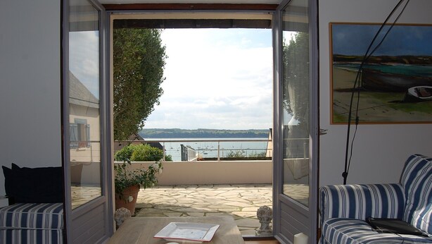 Ein Zimmer mit einer Schiebetür zum Balkon. Der Balkon bietet Blick auf das Meer und Boote. Es gibt ein gestreiftes Sofa und einen kleinen Tisch mit einer Schüssel und einem Buch darauf.