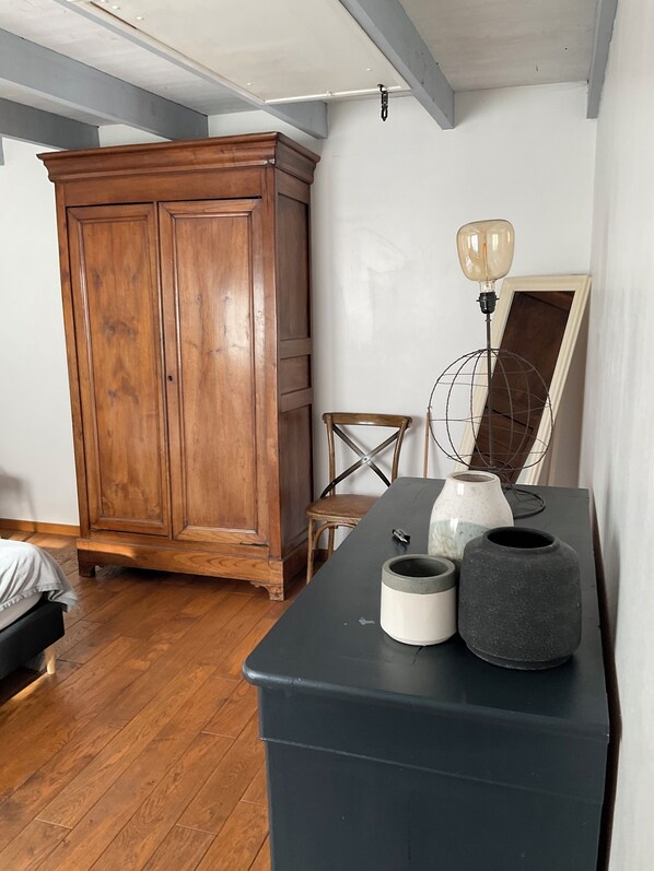 A room with a wooden wardrobe, a chair, a table with decorative items, and a bed.