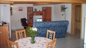 Ein Wohnzimmer mit blauem Sofa, Holzbalkendecke, einem Esstisch mit Stühlen und einer Vase mit Blumen.