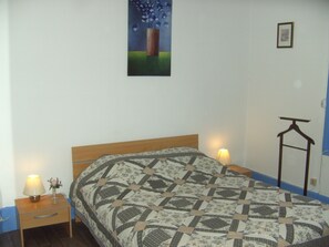 A neatly made bed with a patterned quilt, a bedside table with a lamp and a vase, and a painting on the wall.