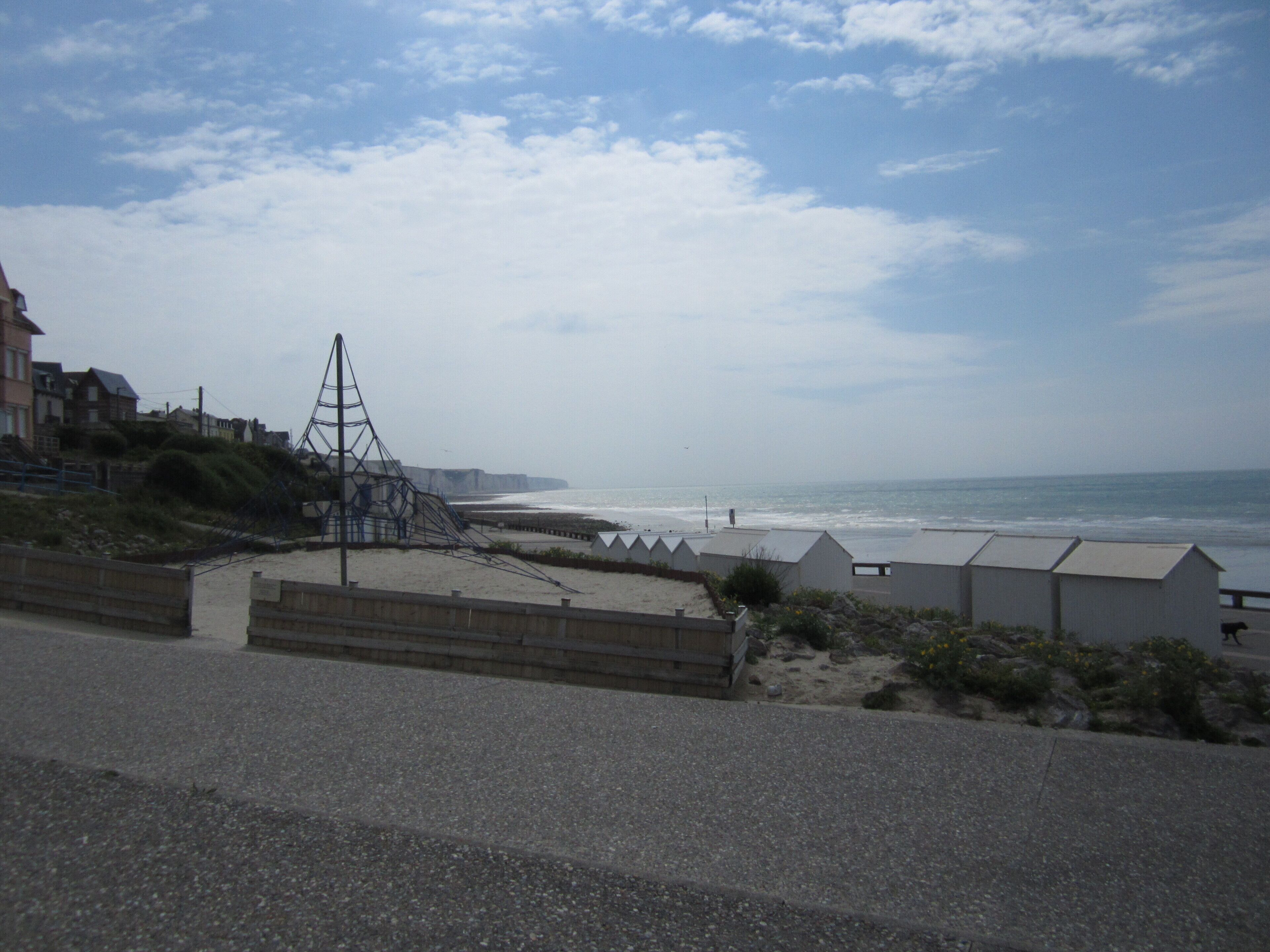 A coastal road with a beach on the right, featuring a large metal structure, a sandy area with small buildings, and a clear sky.