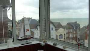 A room with a large window overlooking a coastal town, a small sailboat model, and a decorative lighthouse model on the windowsill. - Apartment in villa for 6 persons-2**- . Sea view, somme bay.    (Ault)