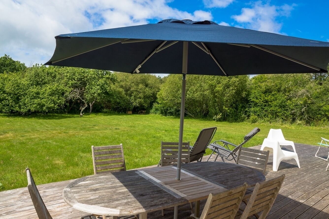 Un patio al aire libre con una mesa y sillas de madera, un paraguas grande y vistas a una zona verde con árboles.