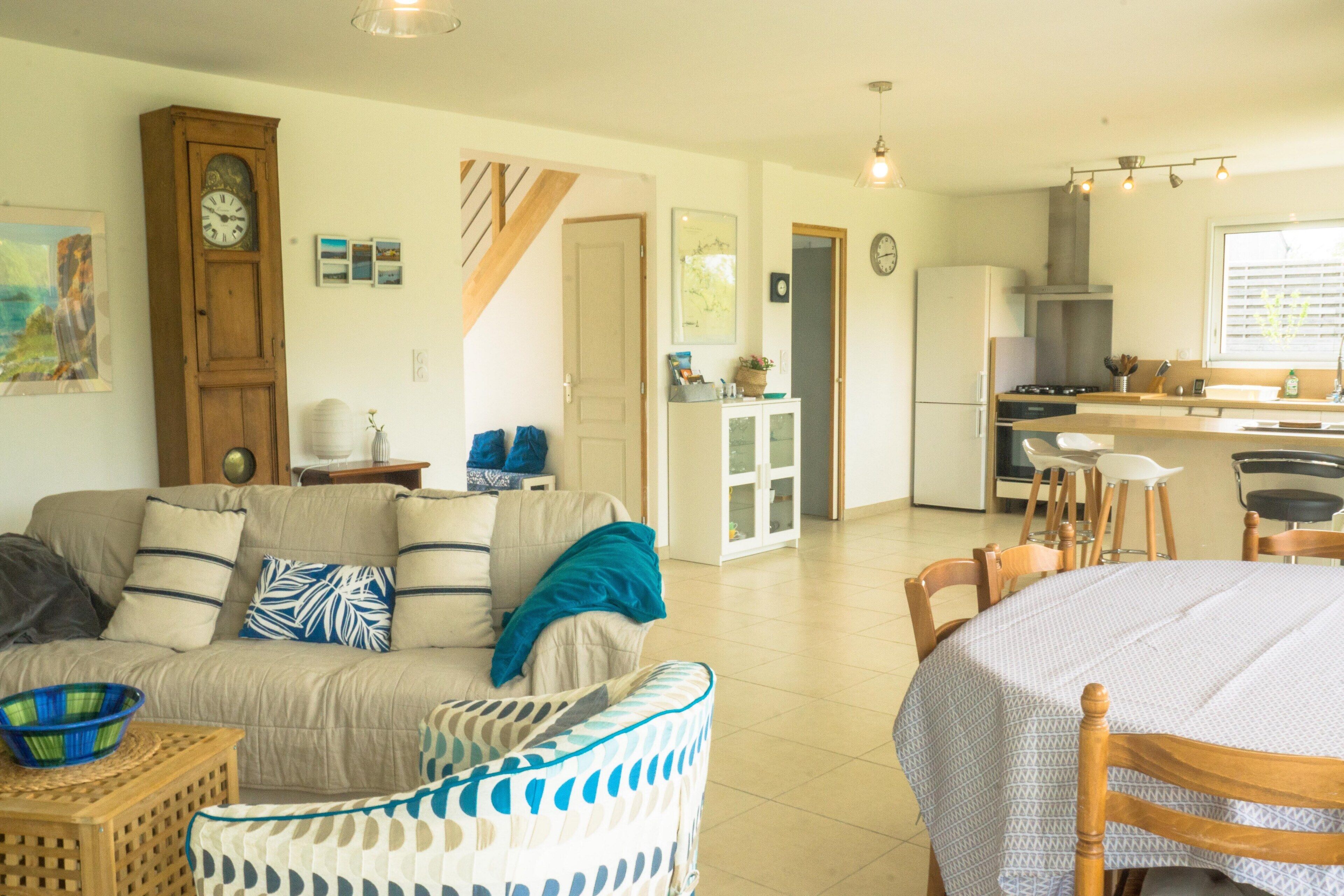Una sala de estar con sofá, una mesa de comedor con sillas, una zona de cocina con refrigerador y una escalera que lleva a otro nivel.