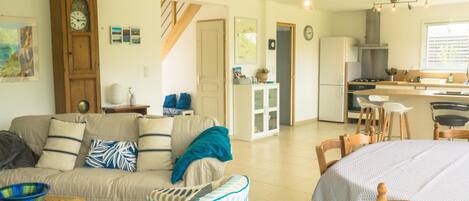 Una sala de estar con sofá, una mesa de comedor con sillas, una zona de cocina con refrigerador y una escalera que lleva a otro nivel.