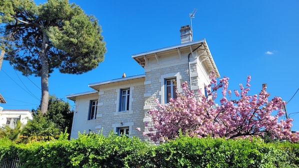 A stone building with a chimney, a large tree, and blooming pink flowers in the foreground.