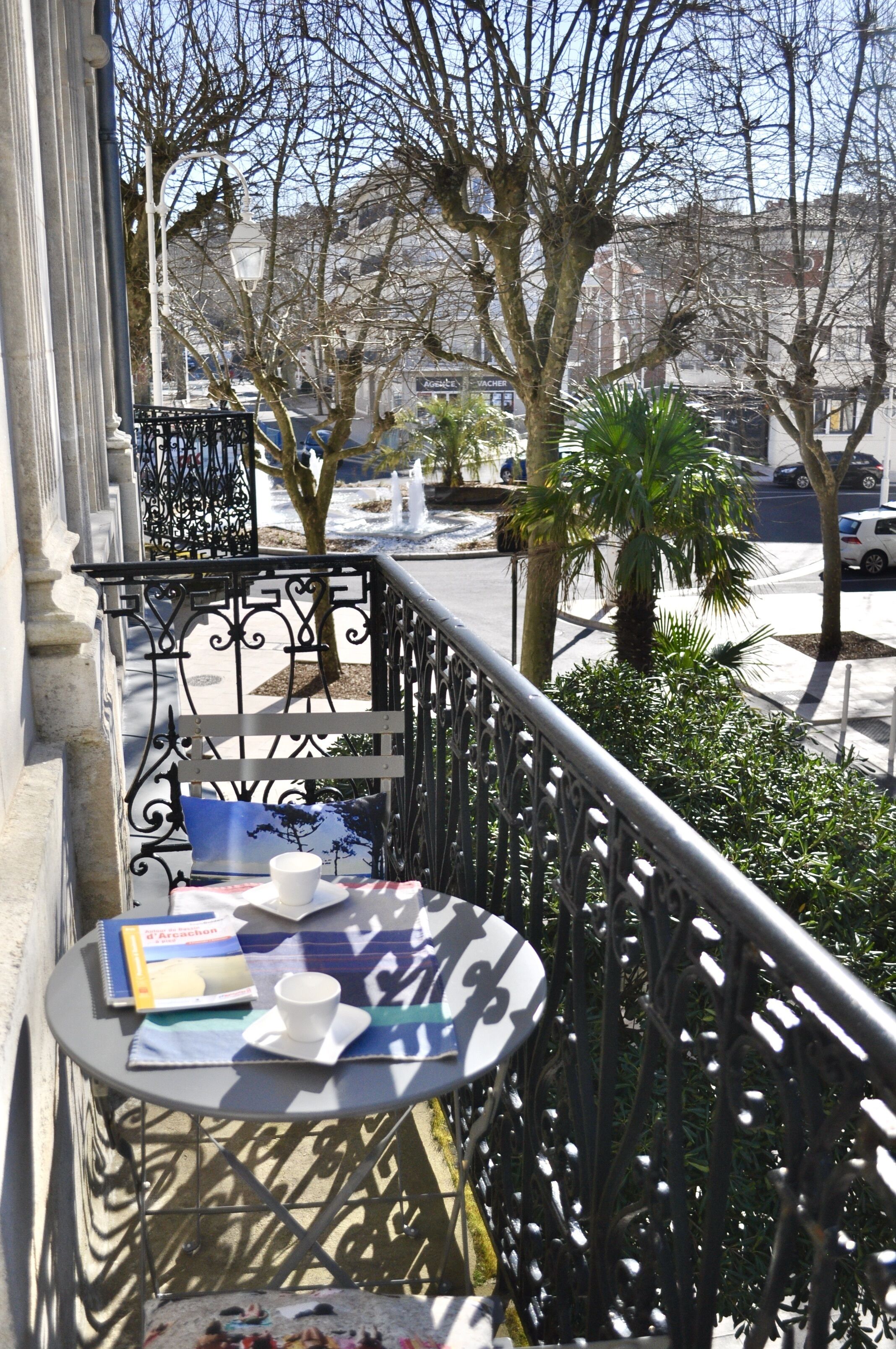 Un balcón con una mesa, una silla y un libro. Se ven árboles y una calle al fondo.