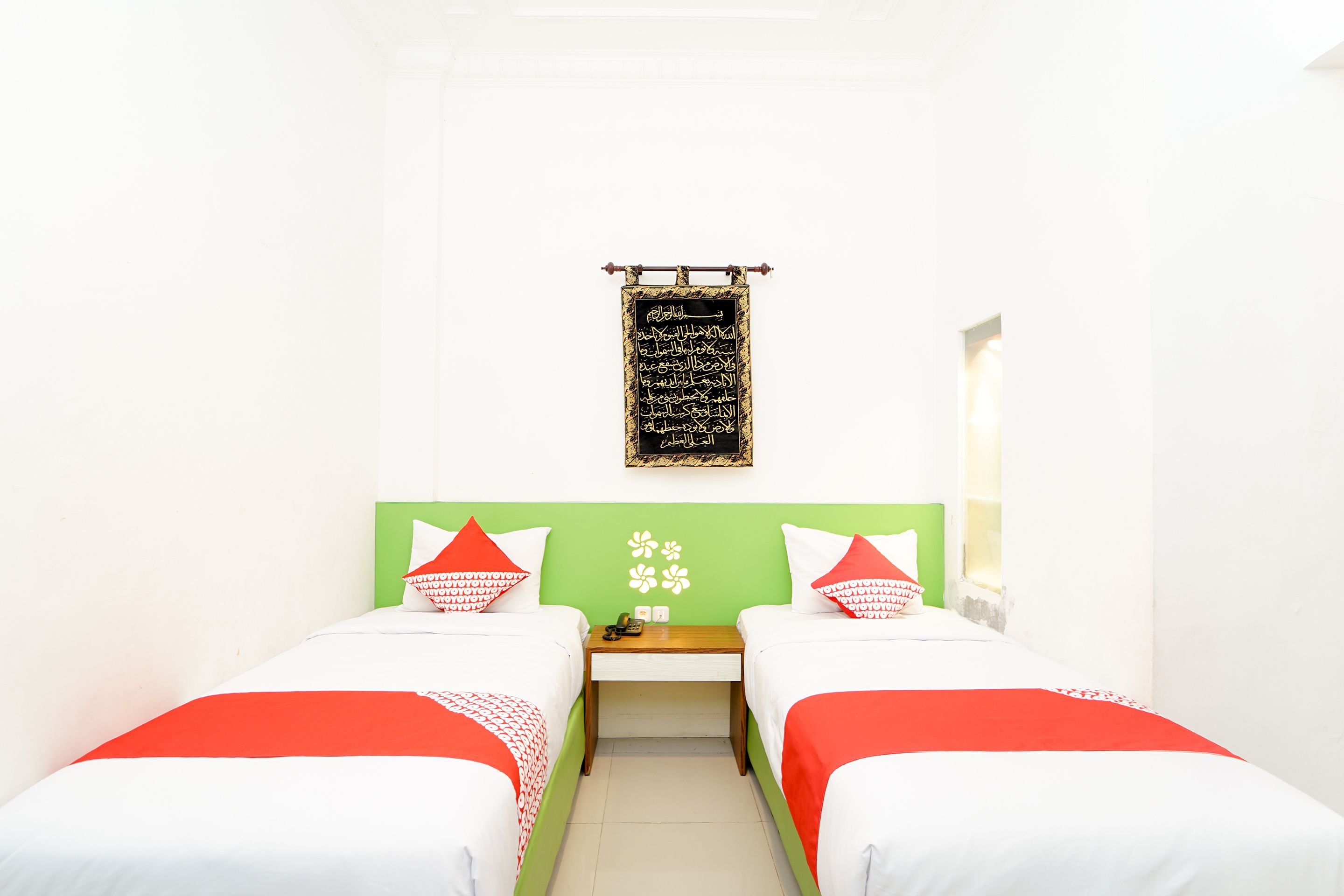 A hotel room with two single beds, a small table, a nightstand, and a wall-mounted blackboard with Arabic calligraphy.