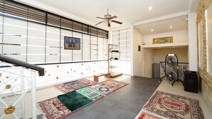 Une pièce spacieuse avec un plafond haut, un ventilateur de plafond et une grande fenêtre avec des stores. Le sol est recouvert de plusieurs tapis et un banc est placé au centre de la pièce.