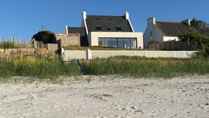 Uma casa moderna com fachada branca, grandes janelas e telhado inclinado, localizada numa praia arenosa com uma cerca branca ao primeiro plano.